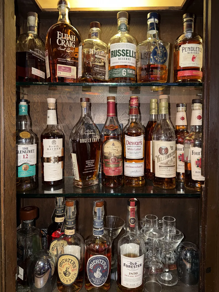 The bourbon cabinet at Grand Crossing: three lit glass shelves of bourbon, rye and scotch including Elijah Craig, Angel's Envy, Michter's and Old Forester 1870