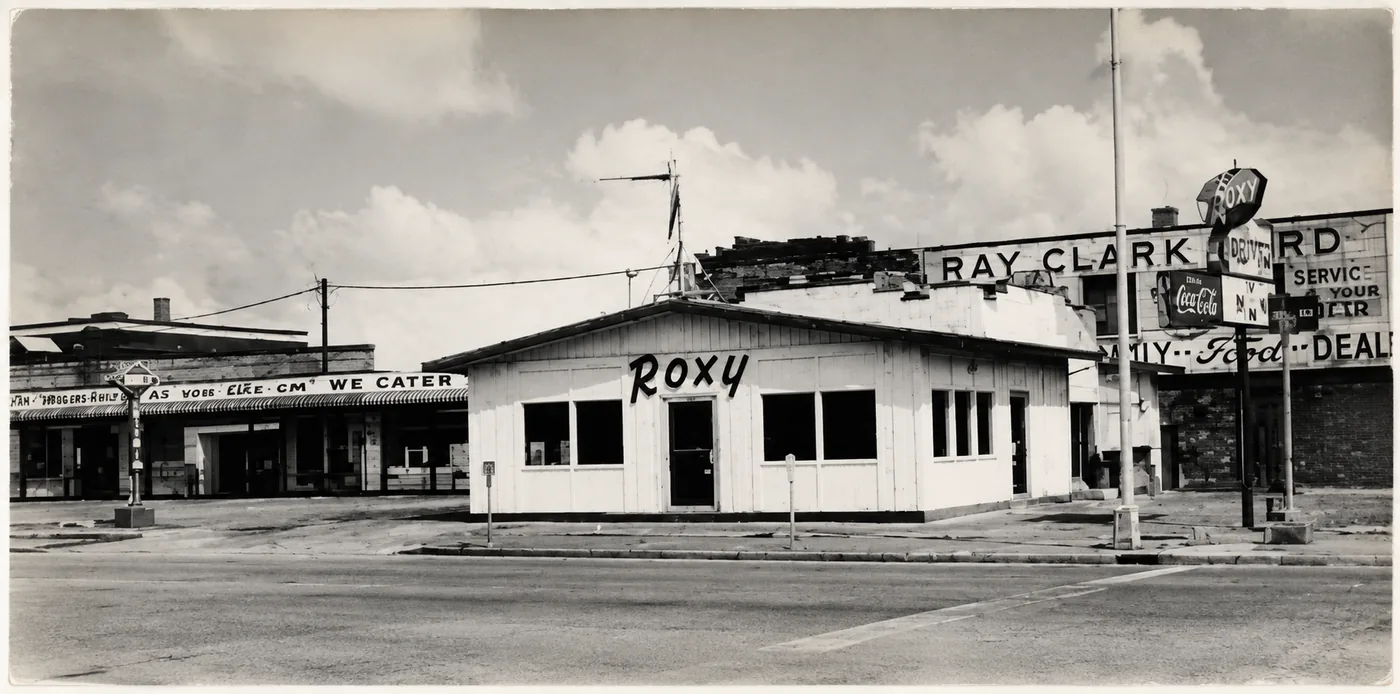 The Roxy, a small white clapboard building with Roxy hand-painted across the side, its drive-in sign on the corner beside a Ford dealership and a Coca-Cola sign